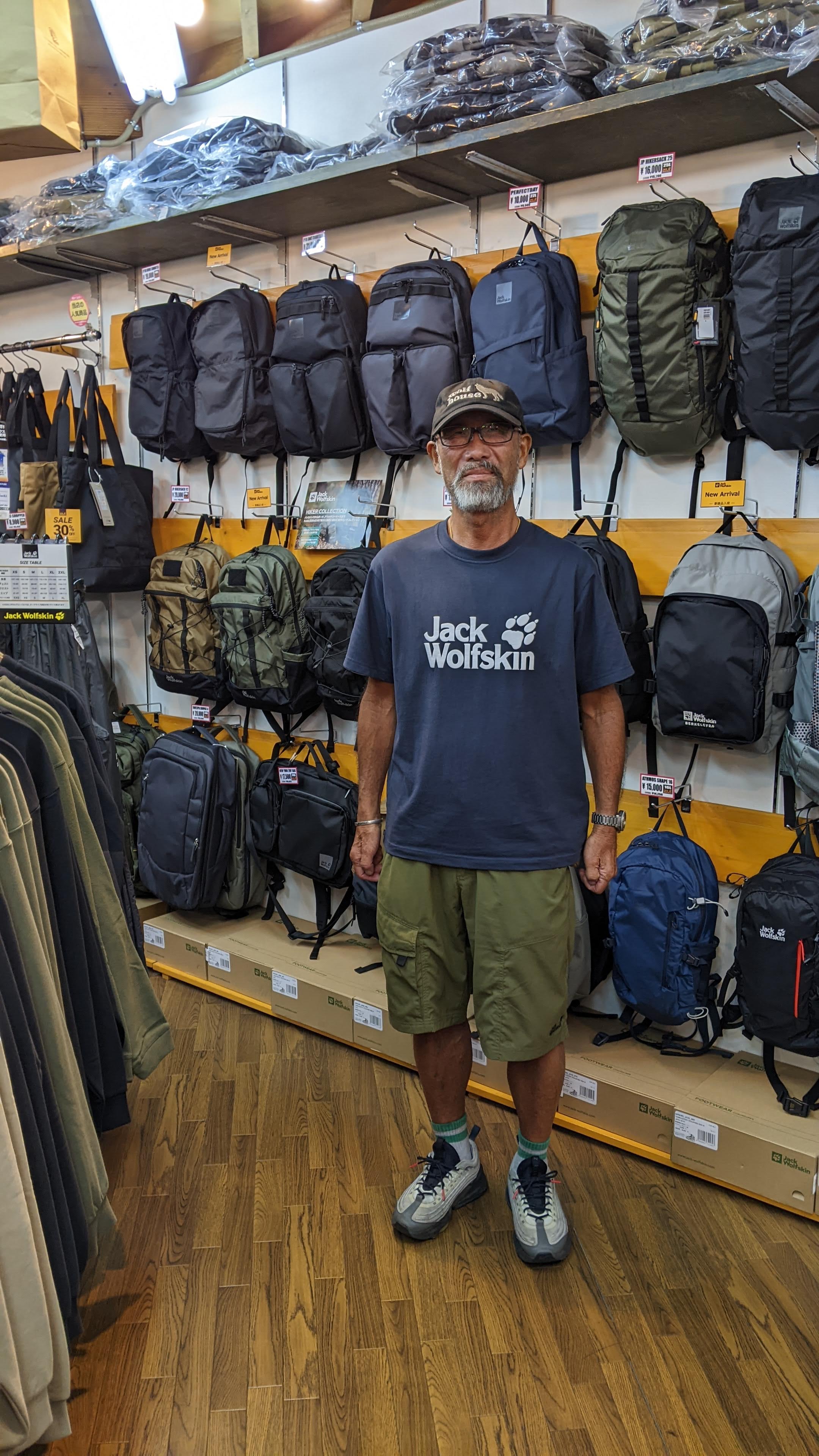 A grey-bearded employee in front of a wall of backpacks